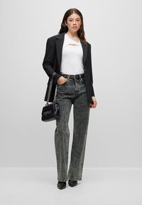 Femme portant un blazer noir, un haut blanc avec une découpe, un jean gris taille haute, des chaussures noires pointues, et tenant un sac à main noir avec une lanière chaîne.