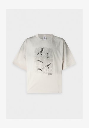 Joven con una camiseta beige con dibujos de figuras corriendo, pantalones negros de pierna ancha y zapatos negros, de pie con una mano detrás de la espalda.