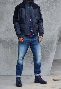 Marinfärgad vadderad jacka med texturerade paneler, utrustad med hög krage och dragkedjor. Matchad med ljusblå denimjeans och marinfärgade kängor.