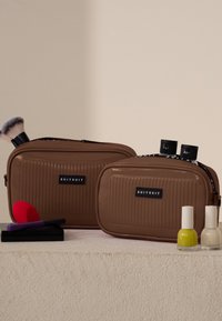 Zwei braune, gerippte Kosmetiktaschen mit Reißverschlüssen und "SUITSUIT"-Etiketten, neben einem Make-up-Pinsel, Nagellack und einem Spachtel.