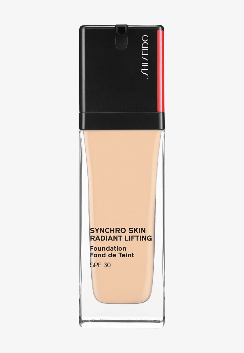 Shiseido Synchro Skin Radiant Lifting -meikkivoiteen pullo mustalla ja punaisella kannella, beige nestemäinen sisältö, näkyvissä SPF 30 -merkintä.