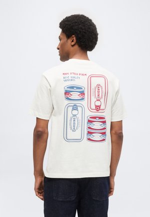 Hombre de espaldas que lleva una camiseta blanca con ilustraciones de latas de sardinas en rojo y azul y texto en la parte trasera.