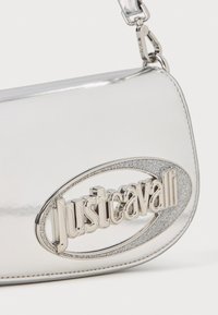 Silver metallic handbag with a shiny Just Cavalli logo plaque and a silver chain strap attached by a clasp in close-up view.