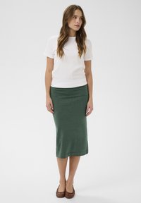 Biely krátky rukáv s rebrovaným vzorom kombinovaný s priliehavou zelenou sukňou po kolená a hnedými tkanými balerínami. Hladká textúra látky a jednoduchý dizajn.