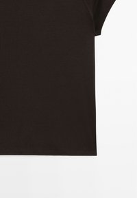 Gros plan sur la moitié inférieure et la couture latérale de la manche d'une chemise à manches courtes brun foncé sur un fond blanc.