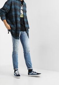 Blue plaid flannel shirt over a black graphic t-shirt, light denim jeans, and navy sneakers with white accents, standing on a light background.