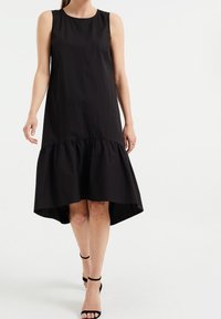 Schwarzes ärmelloses Kleid mit rundem Ausschnitt, das einen rüschengesäumten Saum und einen leicht verlängerten Rücken aufweist. Hergestellt aus glattem Stoff.