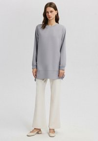 Tunika grigia chiara con maniche lunghe e spacchi laterali, abbinata a pantaloni ampi color crema e scarpe chiare. Tessuto morbido, vestibilità rilassata.