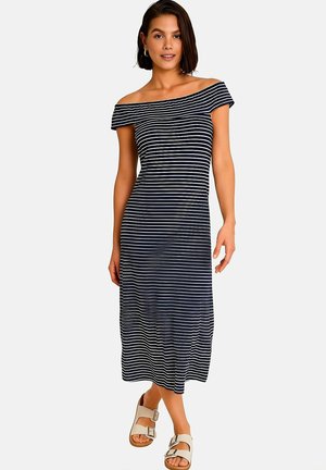 Femme portant une robe midi à rayures bleu marine et blanches, épaules dénudées, avec des sandales beige à boucle, se tenant devant un fond uni.