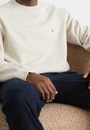 Person trägt einen cremefarbenen langärmeligen Sweatshirt mit kleinem Logo auf der Brust und dunkelblaue Hose, sitzt auf einer beige strukturierten Oberfläche.