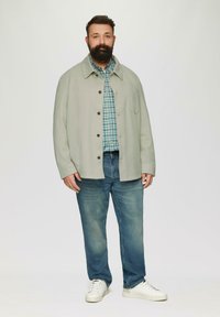 Hellgraue Button-Up-Jacke über einem blauen Karomusterhemd, kombiniert mit blauen Jeans und weißen Sneakers. Glatter Stoff mit lockerer Passform.