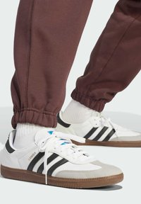 Sneakers in pelle bianca con strisce nere, suola in gomma e dettagli blu. Indossati con pantaloni sportivi color maroon, con polsini elastici.