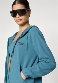 Teal fleece zip-up hoodie with an adjustable hood, side pockets, and a Patagonia logo patch on the left chest. Model wears black sunglasses.