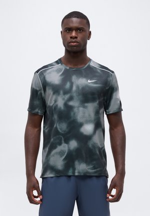 Homme debout portant un t-shirt Nike à manches courtes gris foncé et noir à motifs et un short bleu sur un fond blanc uni.