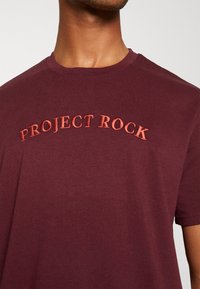 Camiseta de cuello redondo en color burdeos, hecha de una tela suave, con "PROJECT ROCK" bordado en naranja contrastante en el pecho.
