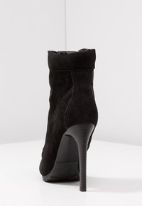 Black suede ankle boot with a sleek, form-fitting design, high stiletto heel, and minimal stitching details. Smooth texture throughout.
