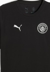 Sort kortærmet skjorte lavet af glat stof. Har et hvidt Puma-logo på venstre bryst og et rundt Manchester City-logo på højre side.