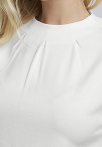 Bílý top s kulatým výstřihem a vertikálními sklady na přední straně, vyrobený z texturované tkaniny. Nabízí hladký, elegantní vzhled.