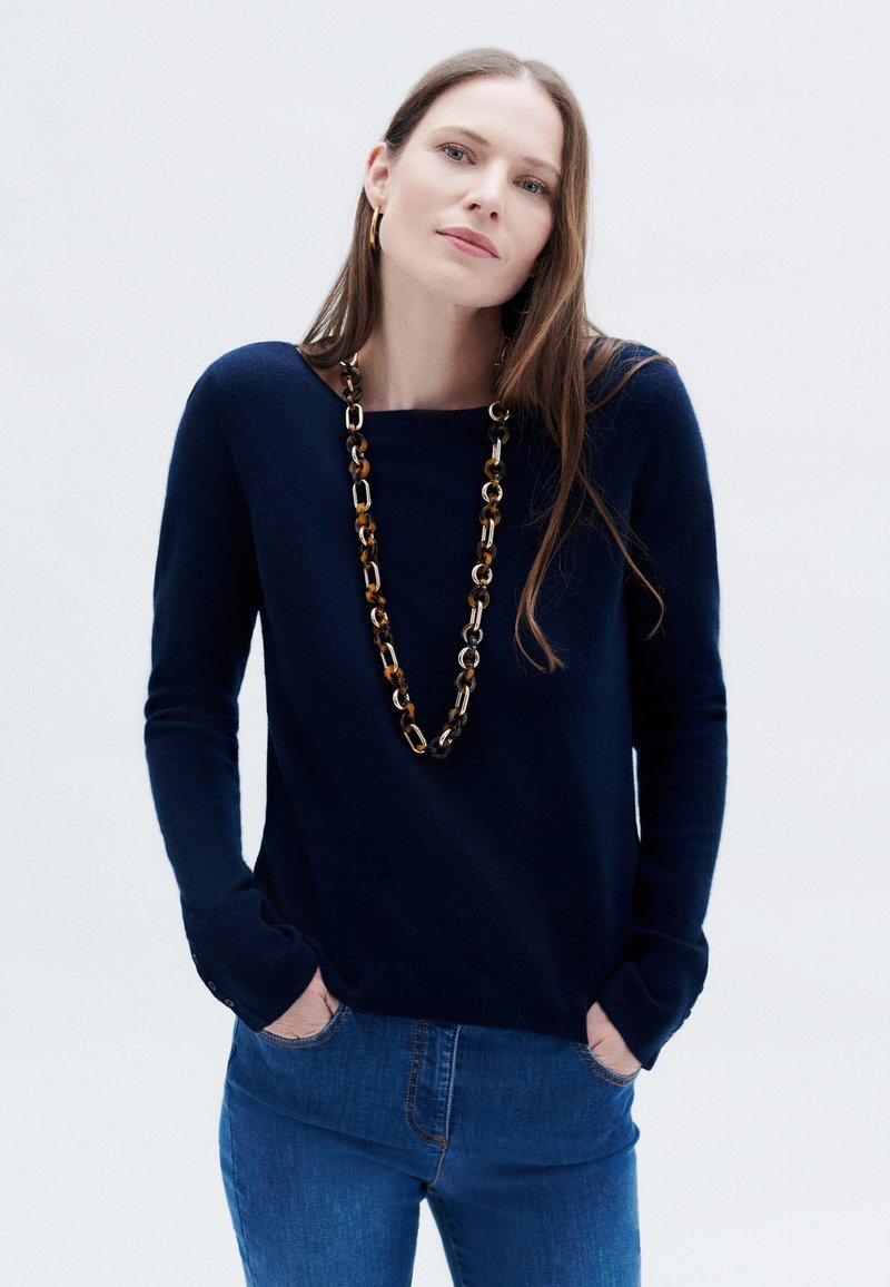 Marineblauer gestrickter Pullover mit langen Ärmeln und einem Bootsausschnitt. Kombiniert mit blauen Jeans und einer langen Kettenhalskette mit ovalen Gliedern.