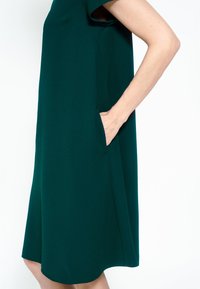 Rochie verde în linie A, realizată dintr-un material uniform, cu mâneci scurte și buzunare laterale. Croiala se termină puțin deasupra genunchiului, cu un tiv rotunjit.