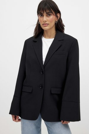 Femme portant un blazer noir surdimensionné avec de larges manches sur un haut blanc et un jean bleu clair, se tenant devant un fond uni.