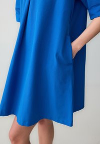 Abito in cotone blu con vestibilità ampia, caratterizzato da maniche corte e tasche laterali. Texture liscia, orlo a ginocchio, senza motivi o accenti visibili.