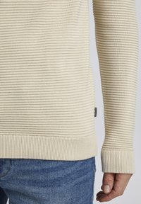 Beige gestreifter Strickpullover mit gerippten Bündchen und Saum, der ein strukturiertes Muster aufweist. Kombiniert mit blauen Jeans.