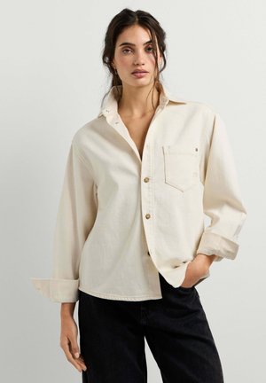 Femme portant une chemise boutonnée écrue aux manches retroussées et un pantalon noir, debout avec une main dans la poche sur fond uni.