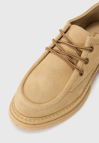 Beige suede shoe with rounded toe, brown laces, and stitched detailing on the sole for added grip and texture.