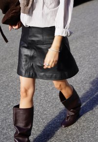 Fekete bőr mini szoknya sima textúrával és egyszerű dizájnnal. Térdmagasságú, laza sötétbarna csizmával párosítva.