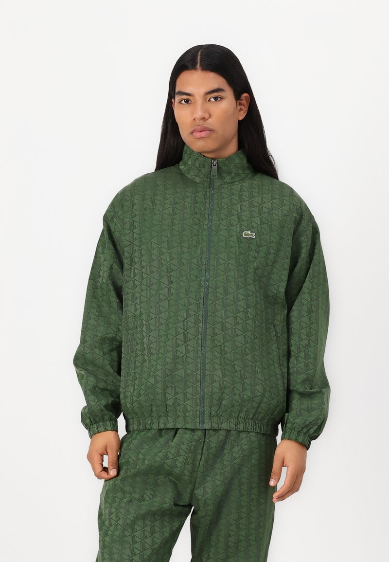 Grüne gemusterte Zip-Up-Jacke mit hohem Kragen, elastischen Bündchen und passender Hose, mit einem dezenten Logo auf der Brust.