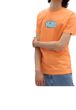 Osoba nosząca pomarańczową koszulkę Vans z niebieskim logo, z założoną na krzyż lewą ręką, częściowo widoczna w jasnoniebieskich dżinsach na białym tle.