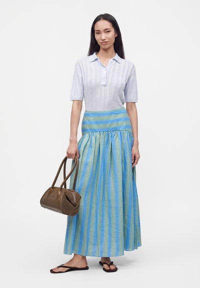 Femme portant une chemise à manches courtes bleu clair et une longue jupe rayée bleu-vert, tenant un sac à main marron et portant des tongs noires.