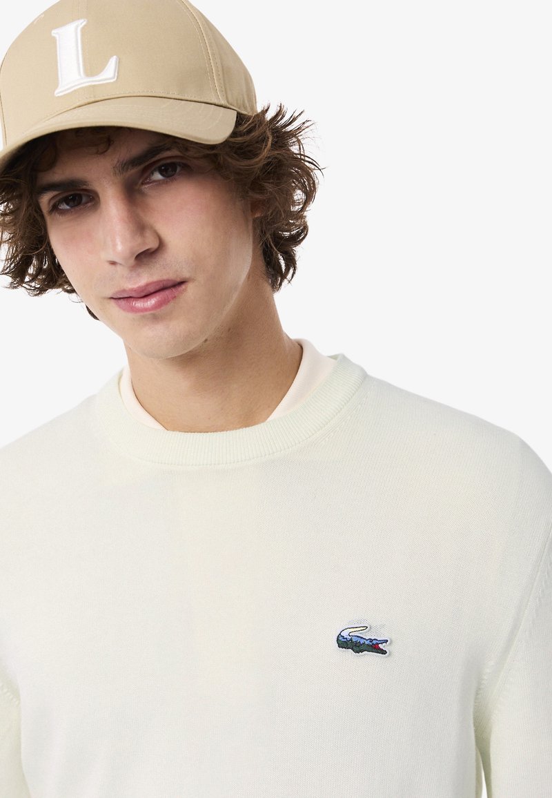 Pull en tricot crème avec un col côtelé, arborant un logo crocodile bleu et vert sur la poitrine gauche. Le modèle porte une casquette beige avec un "L" blanc.