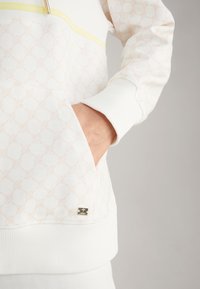 White sweatshirt with a subtle pink geometric pattern, a yellow zipper accent, ribbed cuffs, and a side pocket. Gold logo detail at hem.