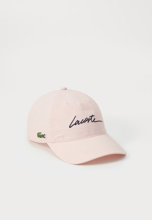 Casquette de baseball rose clair avec le script "Lacoste" noir à l'avant et un petit logo crocodile vert sur le côté, sur fond blanc.