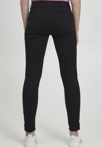 Vaqueros skinny negros hechos de tejido elástico. Cuenta con dos bolsillos traseros, corte ajustado y diseño a la altura del tobillo. Llevados con zapatillas blancas.