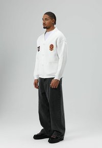 Witte cardigan met knopen aan de voorkant, een bordeauxrood embleem op de borst en het nummer 23. Gecombineerd met donkere oversized jeans en zwarte schoenen.