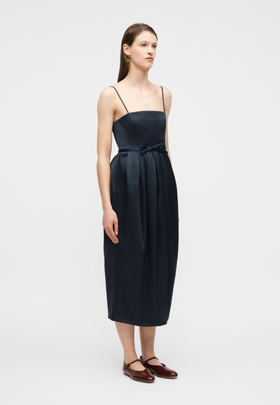 Γυναίκα που φοράει αμάνικο midi φόρεμα σε σκούρο ναυτικό μπλε με λεπτές τιράντες, φιόγκο στη μέση και μπορντό παπούτσια Mary Jane, στέκεται μπροστά σε απλό φόντο.