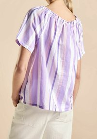 Femme portant un chemisier ample à manches courtes avec des rayures verticales lavande, blanches et roses, assorti à un pantalon beige clair, sur fond beige.