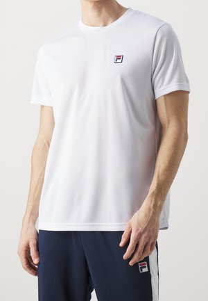 Hombre con camisa blanca de manga corta y pantalones azul marino, ambos con pequeños logotipos de la marca Fila en el pecho y el muslo.
