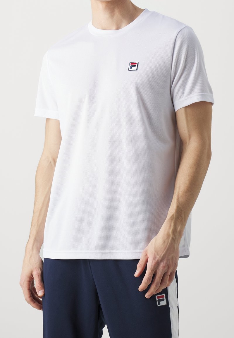 Homme portant une chemise blanche à manches courtes et un pantalon bleu marine, tous deux présentant de petits logos de la marque Fila sur la poitrine et la cuisse.