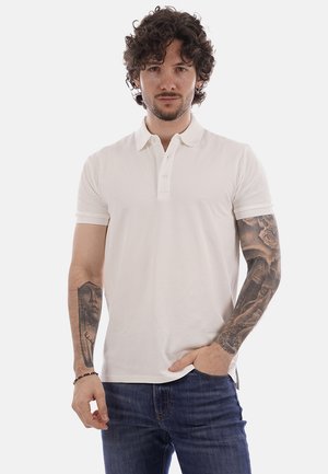 Mann mit lockigem Haar, der ein weißes Poloshirt und blaue Jeans trägt und detaillierte schwarz-graue Tätowierungen an beiden Armen zeigt.