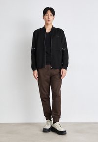 Svart bomberjacka med dragkedja framtill, två fickor och reflexsträcker på ärmarna, ihop med bruna byxor och ljusa sneakers.