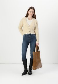Gilet en tricot beige avec des boutons, top blanc, jeans skinny bleu, bottes noires à bout pointu, tenant un sac fourre-tout brillant marron.