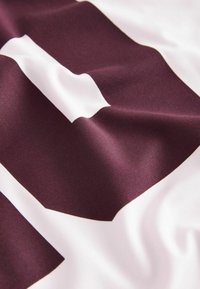 Bordová sportovní dres s bílými akcenty, zdobený výrazným číslem. Měkká, hladká tkanina s viditelnou texturou a jemným leskem.