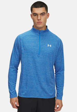 Long-sleeve pullover in textured blue fabric with a quarter zipper, featuring a white logo on the chest and athletic fit.