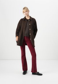 Cappotto button-up marrone scuro sopra un maglione grigio a trecce, abbinato a pantaloni a campana in velluto rosso e scarpe nere. Design semplice e strutturato.