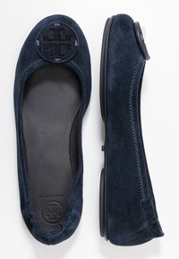 Ballerines en suede bleu marine avec un logo rond à l'avant, intérieur noir lisse et semelle en caoutchouc flexible. Présente des détails de couture contrastants.