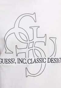 T-shirt bianco con un design grafico nero caratterizzato da lettere sovrapposte e il testo "GUESS?, INC. CLASSIC DESIGN" sottostante.